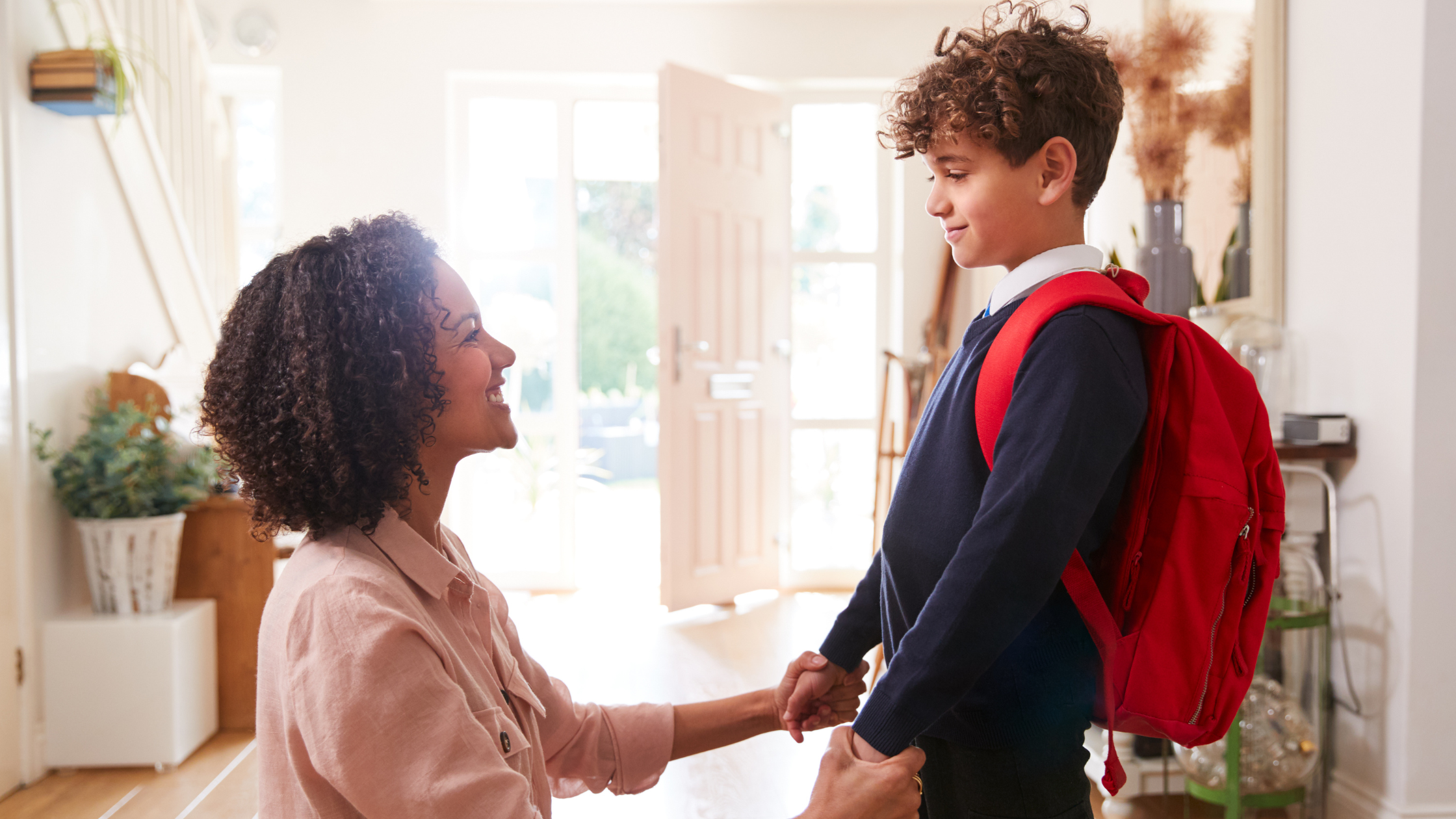 A mother and son holding hands at home, with the boy wearing a backpack.