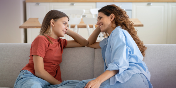 Mom and daughter sit close together on a gray sofa in a bright kitchen, smiling and talking with their hands near their faces, suggesting a friendly conversation.